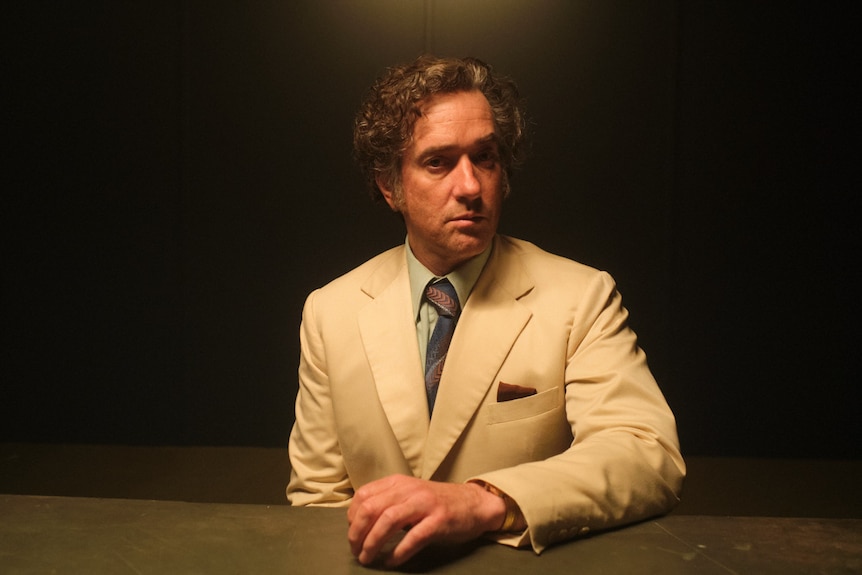 A male actor with brown curly hair poses for a photo in a beige suit and dark striped tie, against a black background