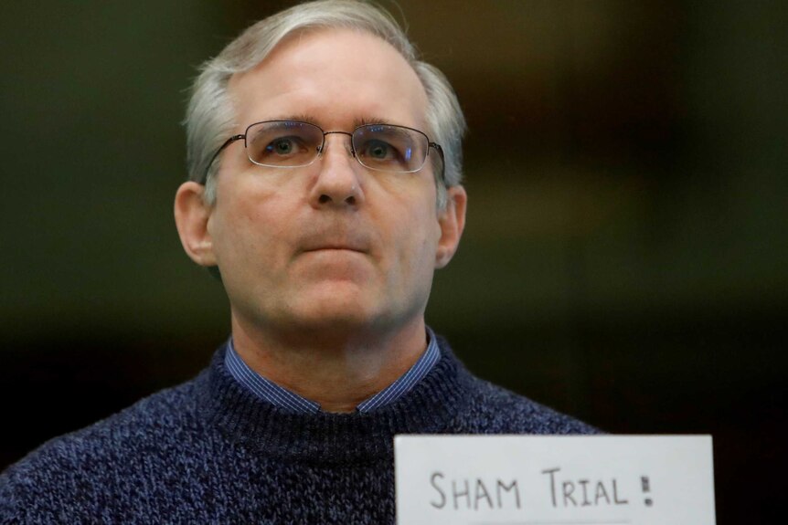 A man wearing glasses standing with a sign which reads 'sham trial!'.
