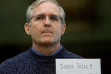 A man wearing glasses standing with a sign which reads 'sham trial!'.