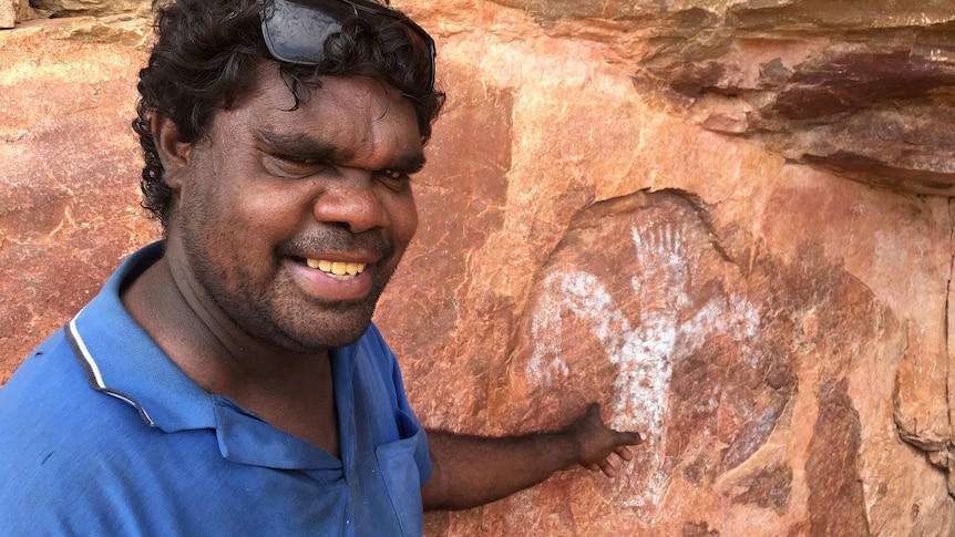 Wesley Alberts stands smiling and pointing at an Indigenous rock art figure.