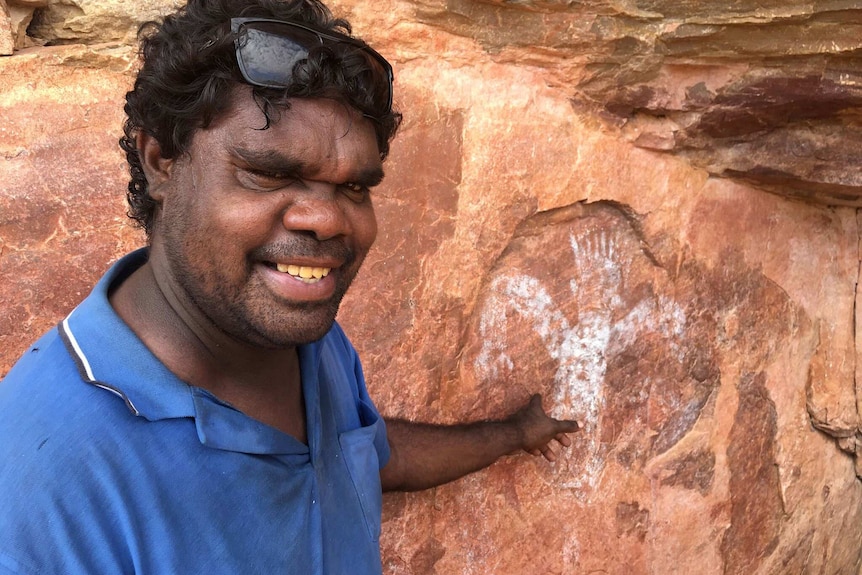 Wesley Alberts stands smiling and pointing at an Indigenous rock art figure.