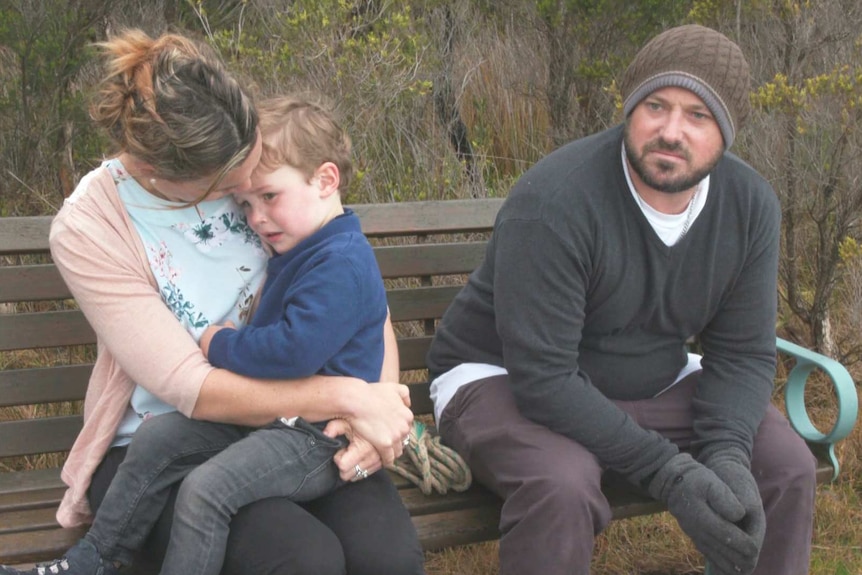 Braden Barnes sits on a bench in the bush. His wife Renee sits on his right with one of their sons sitting on her knee