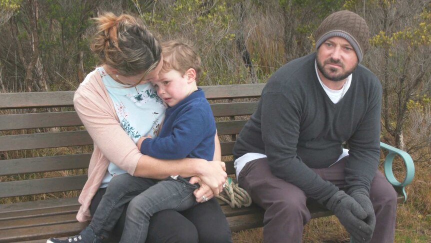 Braden Barnes sits on a bench in the bush. His wife Renee sits on his right with one of their sons sitting on her knee