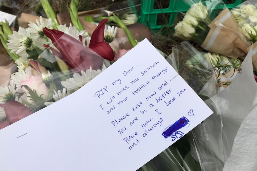 A card reads 'RIP my Dear...I will miss you so much and your positive energy' laid on top of a bunch of flowers.