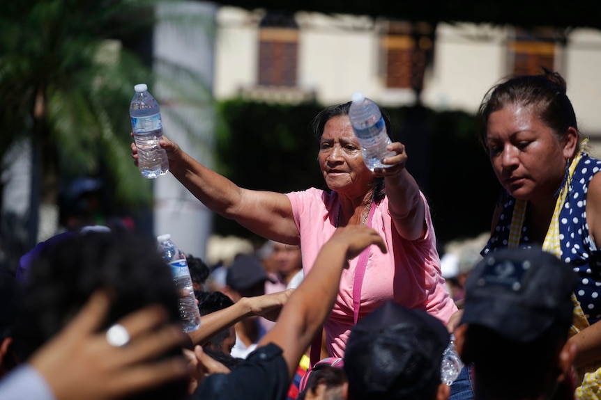 A woman in a pink t-shirt and another in a blue and white spotted shirt hand out water bottles as numerous hands reach for them