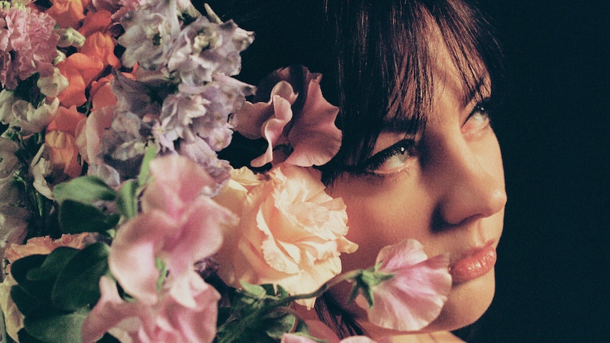 A close-up of Angel Olsen looking wistfully into the distance but the left side of her face is obscure by a bouquet of flowers