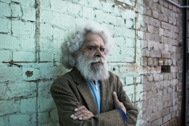 Uncle Jack Charles leans against a brick wall with his arms crossed
