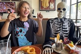 A teenage girl with brown hair sits next to a skeleton in a restaurant with her thumbs up.