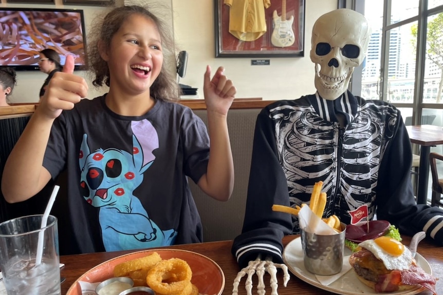 A teenage girl with brown hair sits next to a skeleton in a restaurant with her thumbs up.