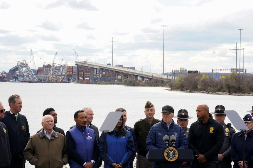 Joe Biden speaks at a press conference with the partially collapsed bridge seen in the background.