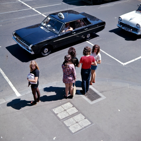 A black taxi passes people dressed in 1960s fashion