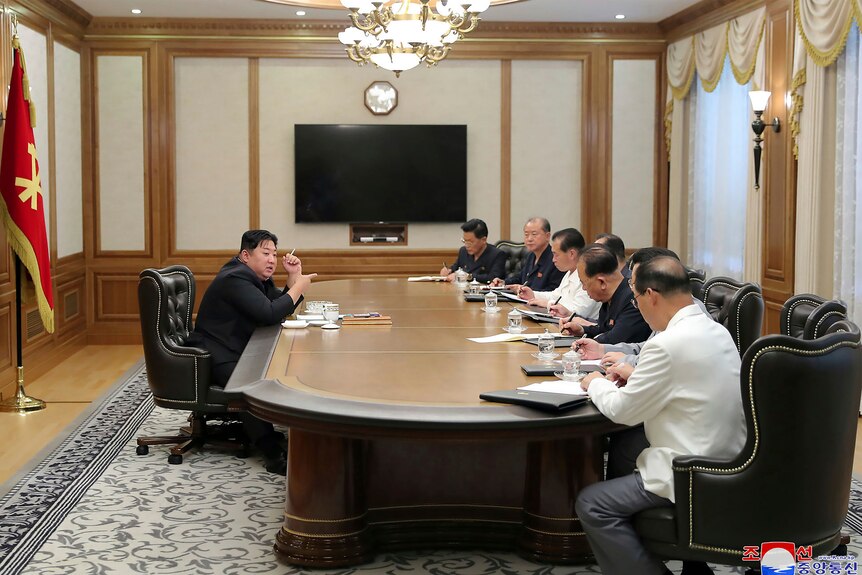 Kim Jong Un sits on one side of a desk, speaking to a group of people on the other side