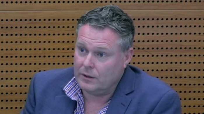 A man wearing a suit jacket sits in the witness box at the royal commission in an image taken from a webstream of a hearing.