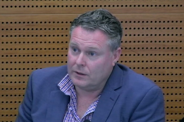 A man wearing a suit jacket sits in the witness box at the royal commission in an image taken from a webstream of a hearing.