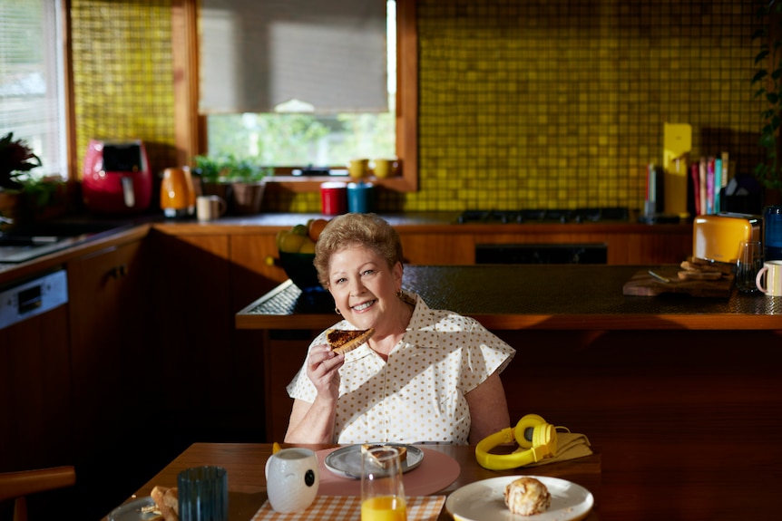 Older lady sits in kitchen holding a piece of vegemite toast and smiles at the camera.