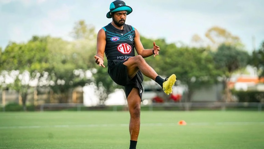A man in teal and black football guernsey and hat kicking on a field.