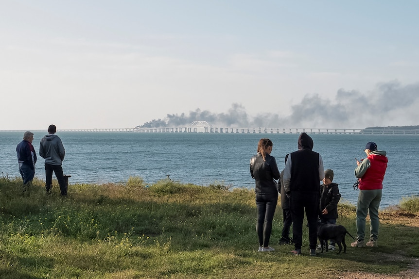 People watch fuel tanks ablaze and damaged sections of a bridge. 