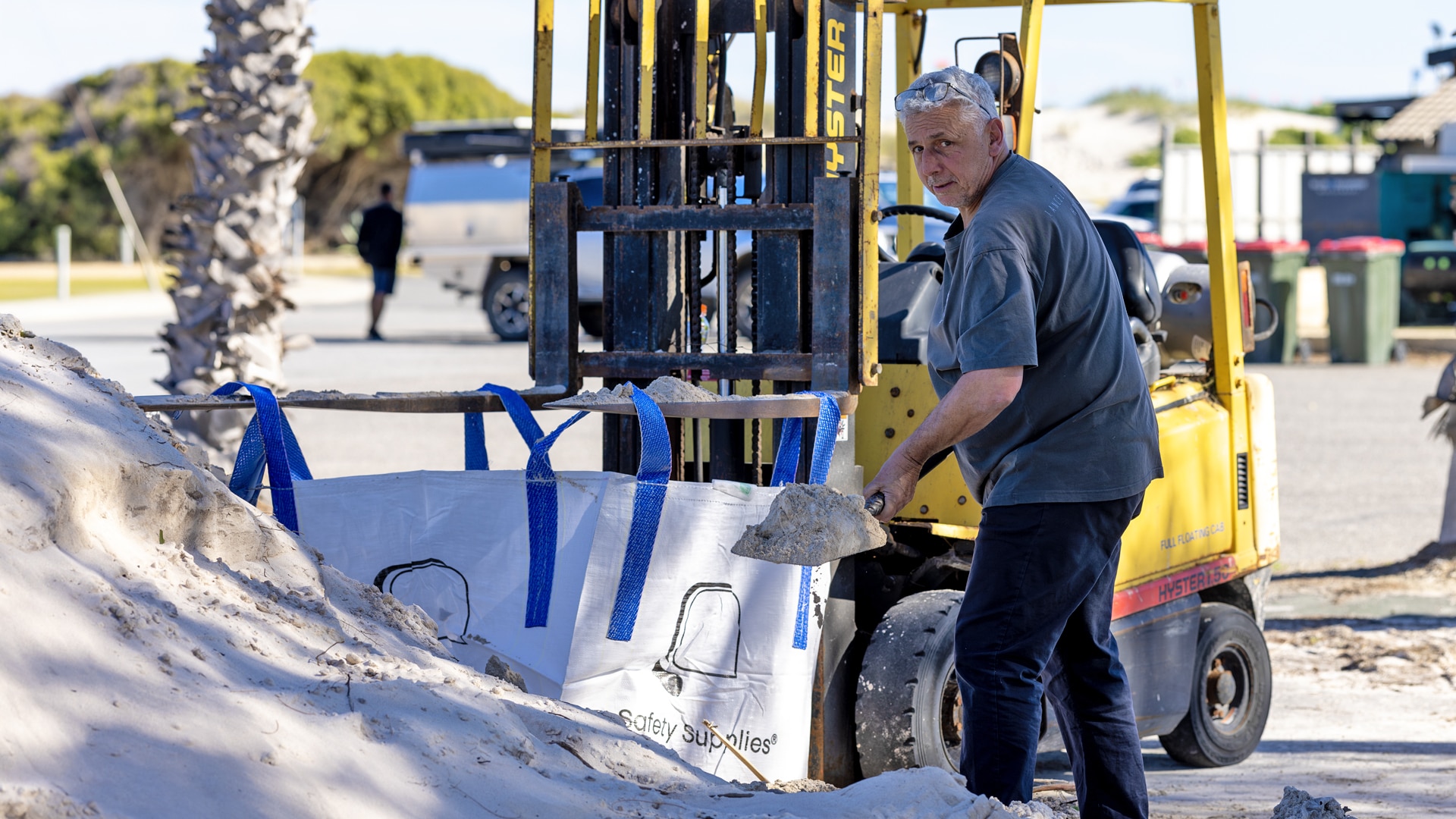 Glen Trebilcock lifts a shovel full of sand, in front of a fork lift holding an open sand bag 