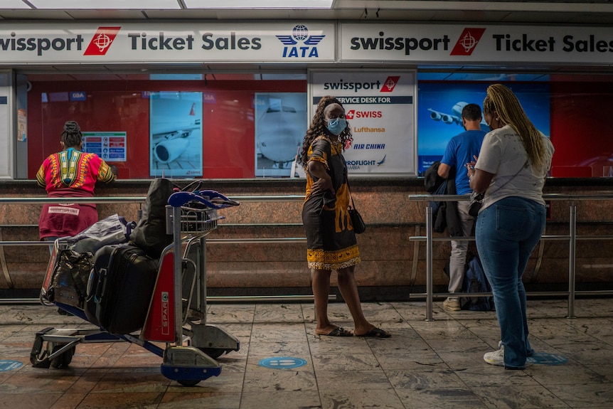 Una mujer de cabello castaño oscuro con un vestido se encuentra con la mano en la cadera en un mostrador del aeropuerto con un carrito cerca