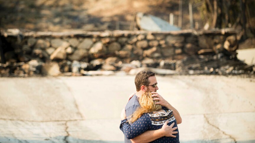 A couple hug in front of the out-of-focus ruins of their home.