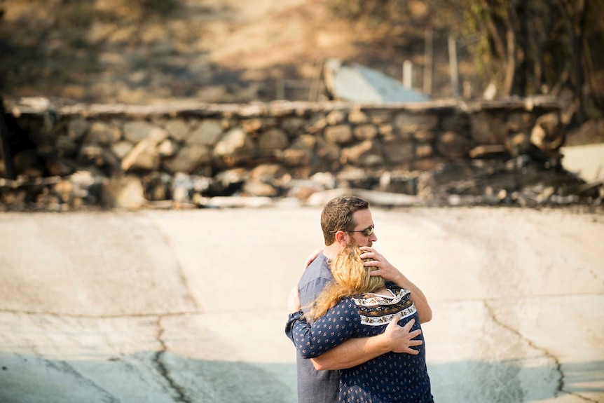 A couple hug in front of the out-of-focus ruins of their home.