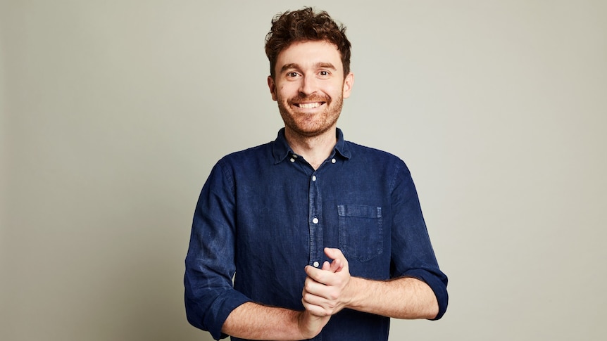 Young man with brown hair, in a blue denim shirt and with clasped hands, smiles at the camera,