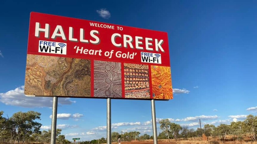 A red road sign with the words Halls Creek, 'Heart of Gold' Free Wi-Fi, Indigenous motives against a blue sky, red earth, trees.