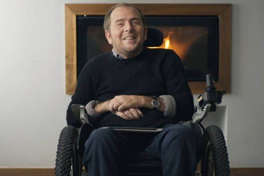 David sits in his wheelchair and smiles as he speaks in a living room in front of a fire.