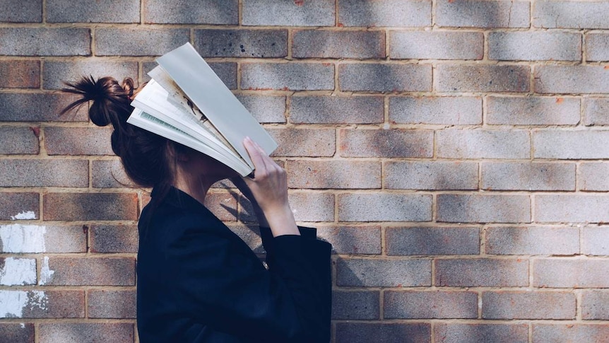 A woman stand in front of a brick wall, holding a textbook over her face