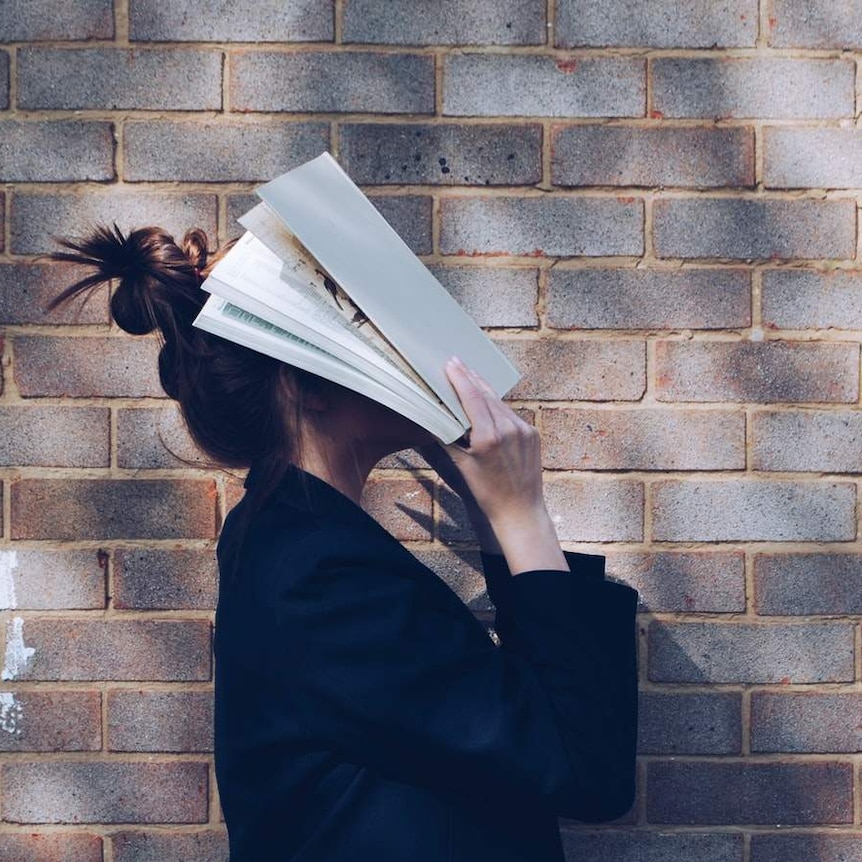 A woman stand in front of a brick wall, holding a textbook over her face