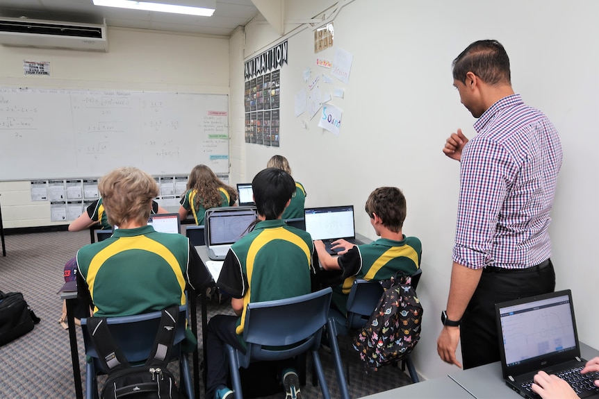 Teacher stands behind a group of students in green and yellow uniforms sitting at a table working.