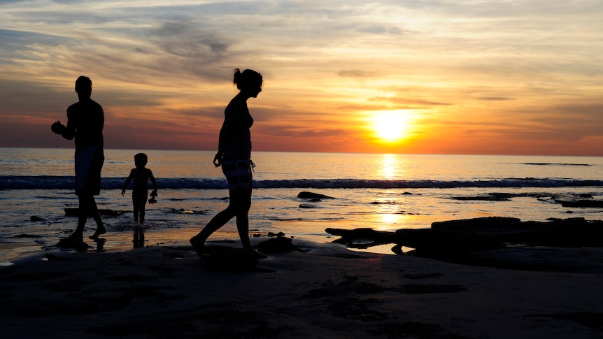A family of three walk along a beach to the backdrop of a sunset