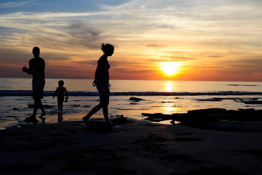 A family of three walk along a beach to the backdrop of a sunset