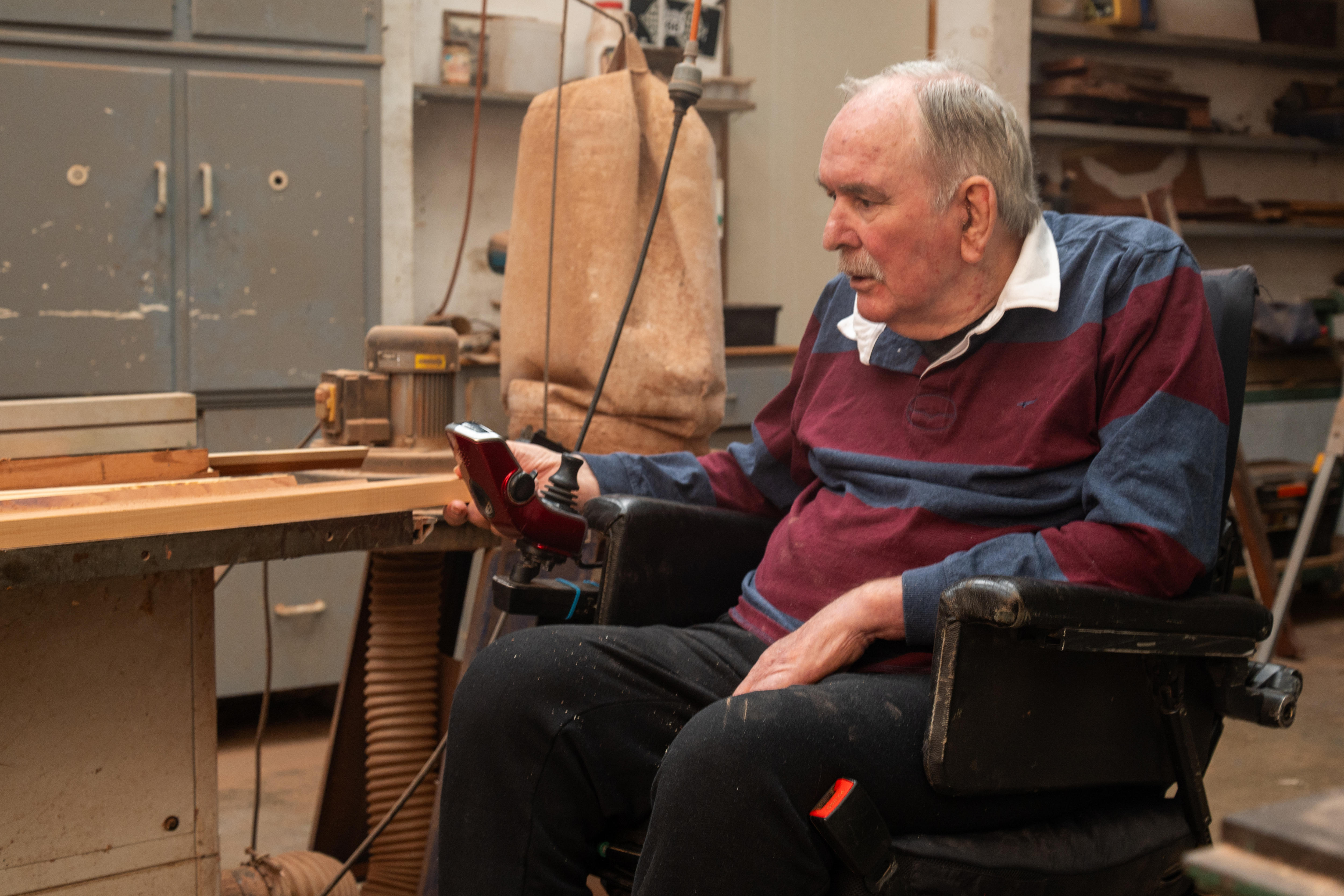A man in a motorised wheelchair holding a small plank of timber in a workshop