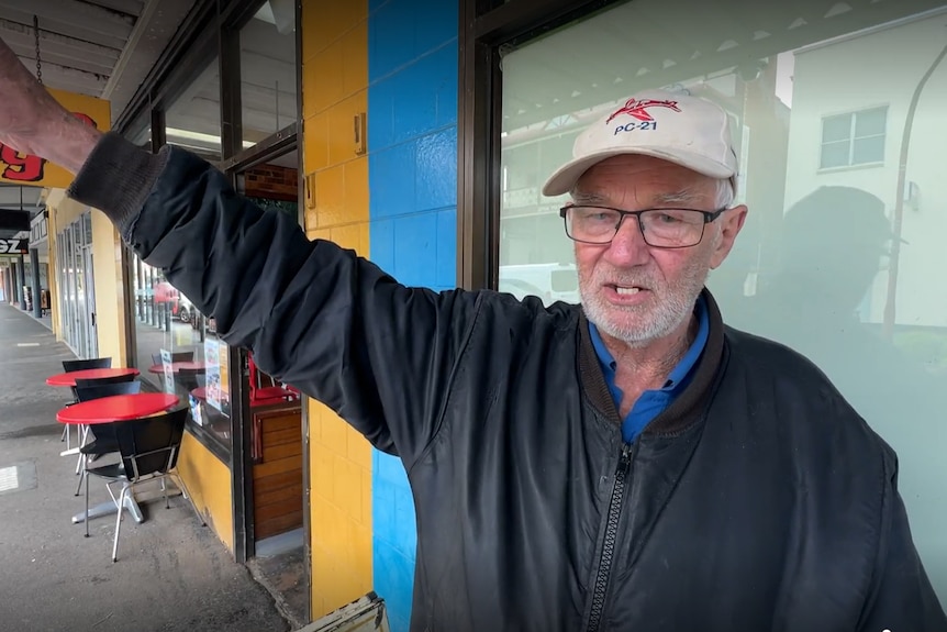 An elderly man wearing a cap points into thin air. He is on an uncrowded main street. it is wet.