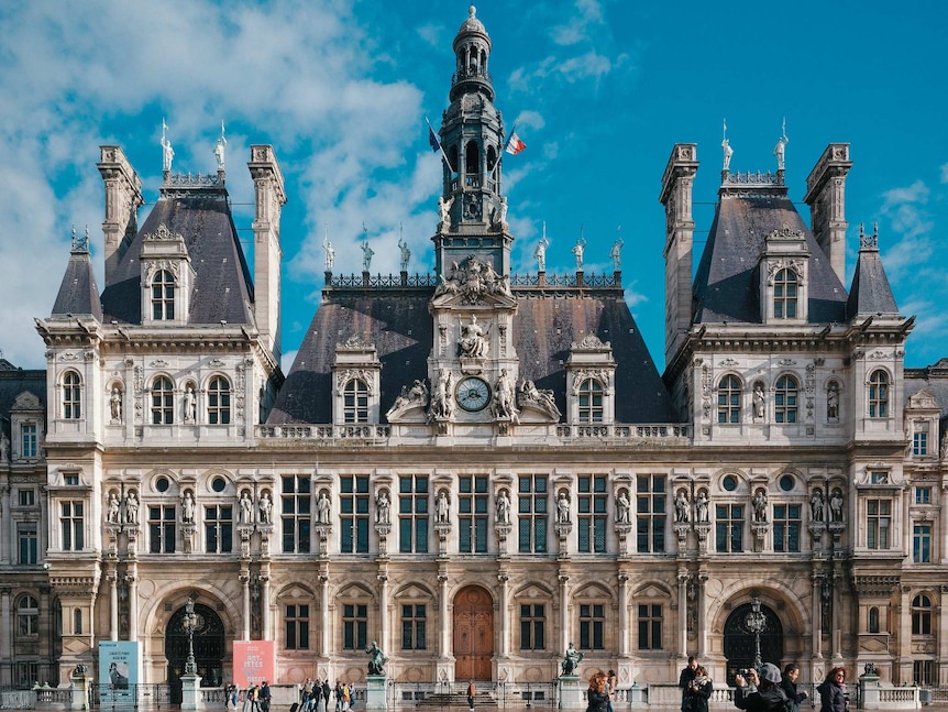 A grand Parisian classical building is pictured front-on on a clear day, with pedestrians in the forecourt.