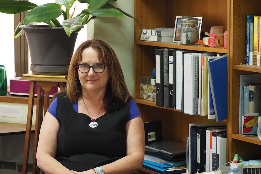 A woman in glasses sits at a desk with a bookshelf behind her.