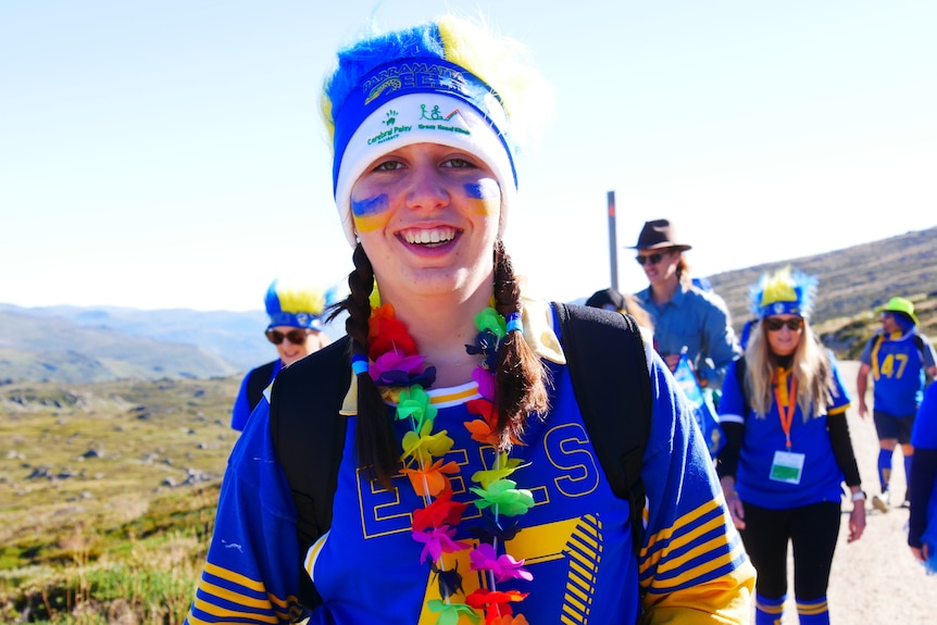 a young girl smiles at the camera whilst climbing a mountain, wearing bright blue colours
