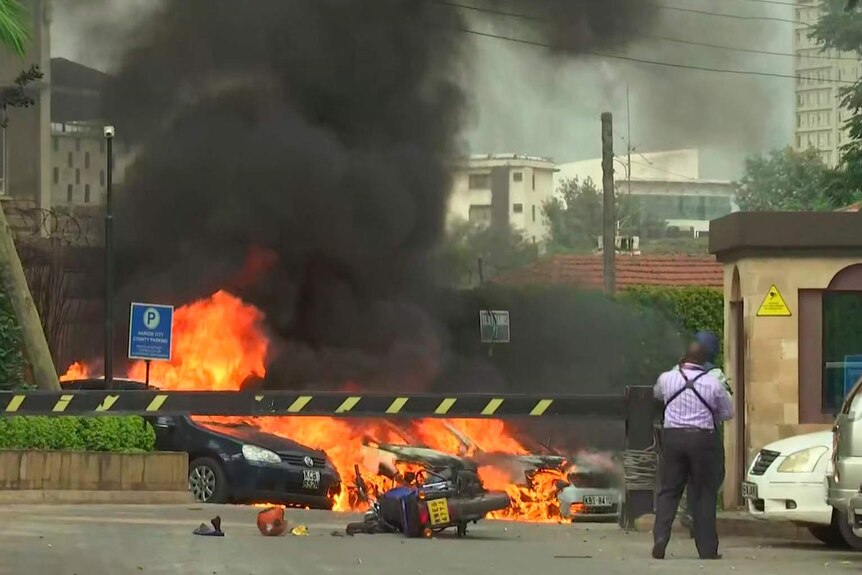 Flames rise from vehicles behind a boom gate as men with guns look on.