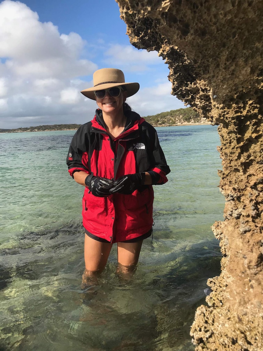 A woman stands in knee deep water wearing a red parka, sun glasses and a wide-brim hat.