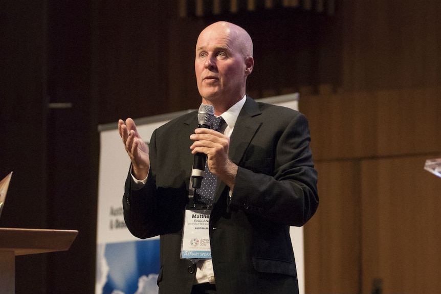 A bald man wearing a dark jacket holds a microphone.