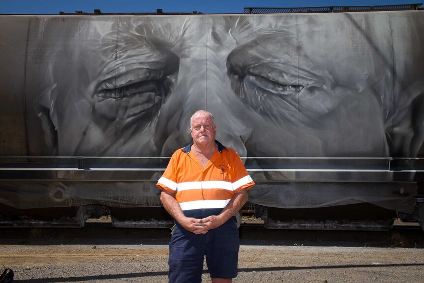 A man in high visibility factory gear standing in front of a train carriage painted with his portrait