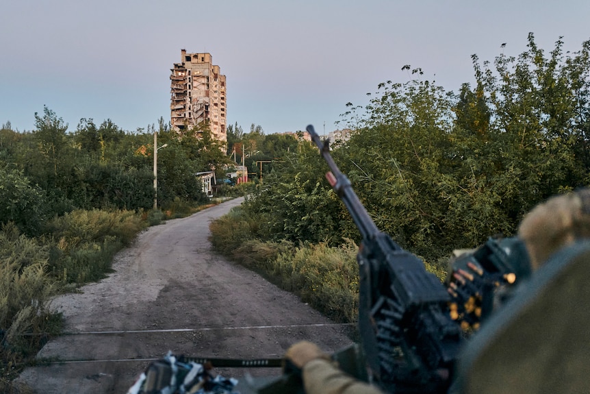 A soldier aims a large machine gun down a rural road with a damaged apartment building in the distance.