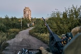 A soldier aims a large machine gun down a rural road with a damaged apartment building in the distance.