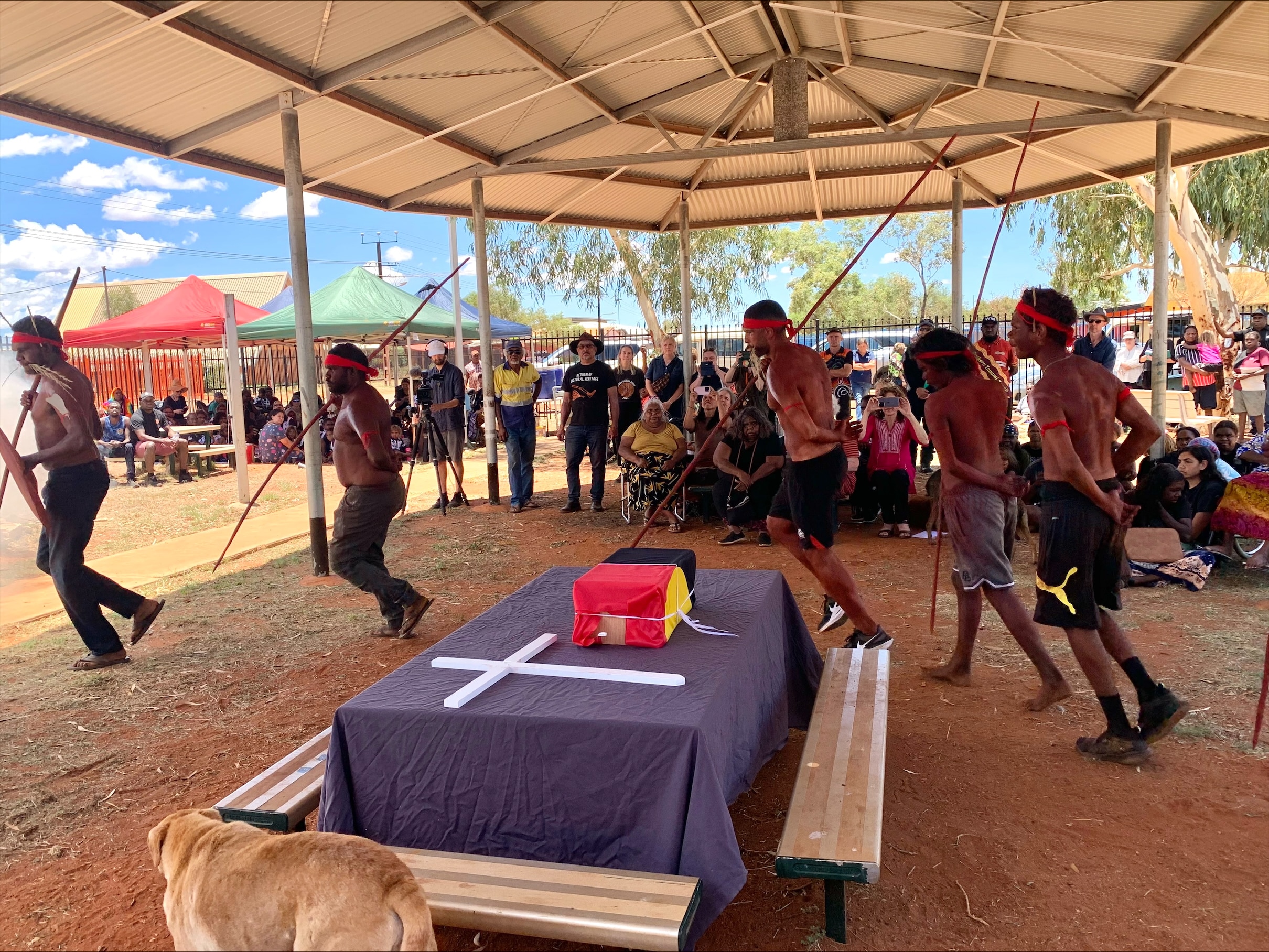 Aboriginal men with spears dance around a table with a cross on it and a box of ashes