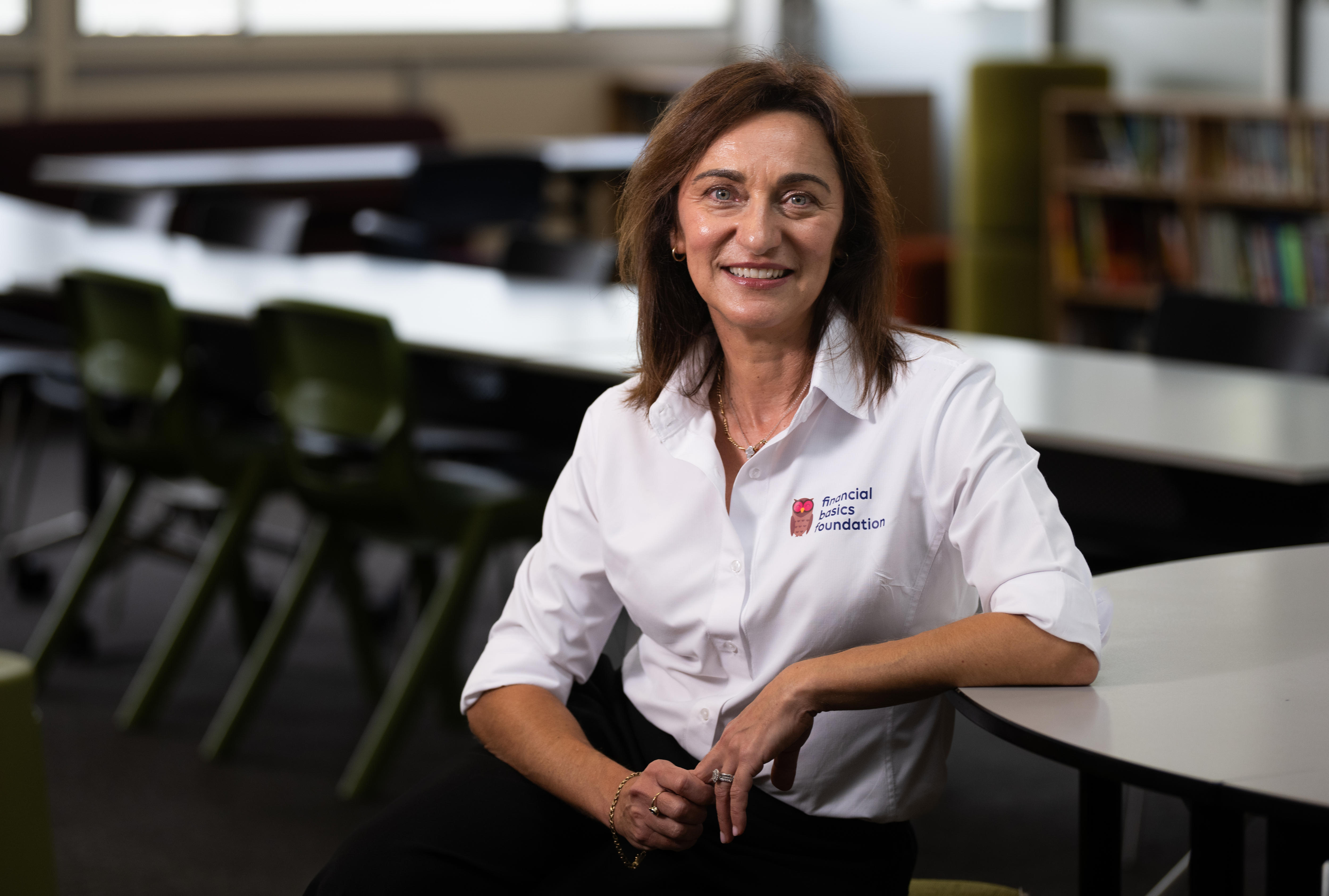 Una mujer de cabello castaño liso, vestida con una camisa blanca de longitud media, sonríe y apoya el codo en un escritorio.