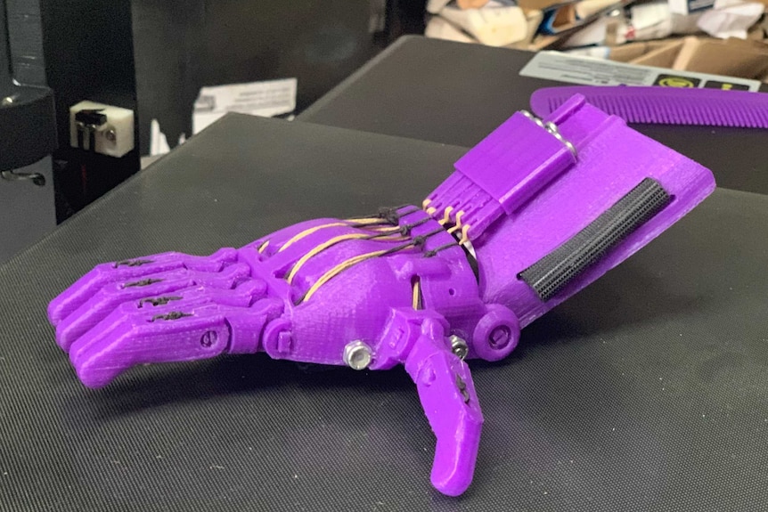 A purple 3D printed prosthetic hand made from recycled plastic  sits on a 3D printer