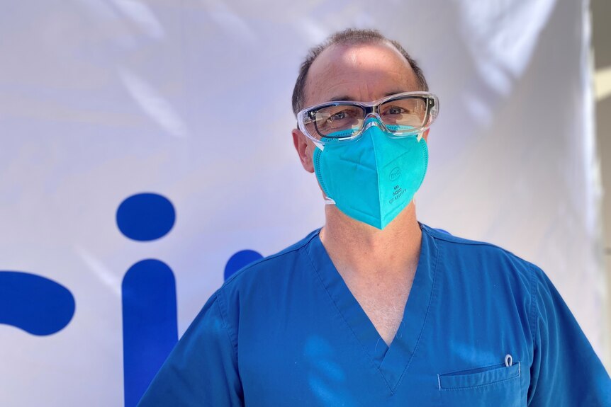 A man wearing blue medical scrubs, a mask and eye shield, standing outside a vaccination clinic.