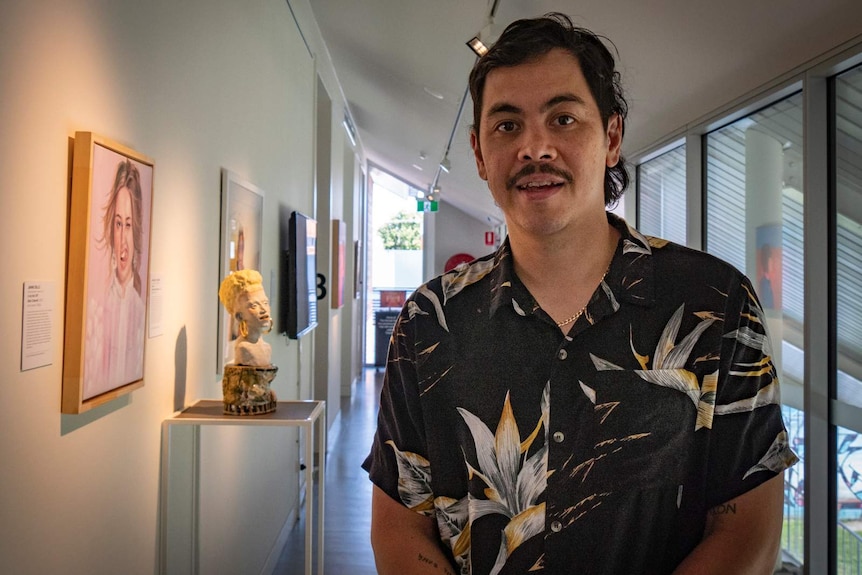 Man on right stand in gallery room with paintings and a ceramic bust on a stand behind him.