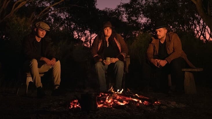 Two men and one woman are sitting around a campfire at dusk. 